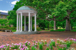 Surrounded by ancient oak trees, lush lawn and flowers, the iconic Old Well at the University of North Carolina Chapel Hill is pictured with blue skies.