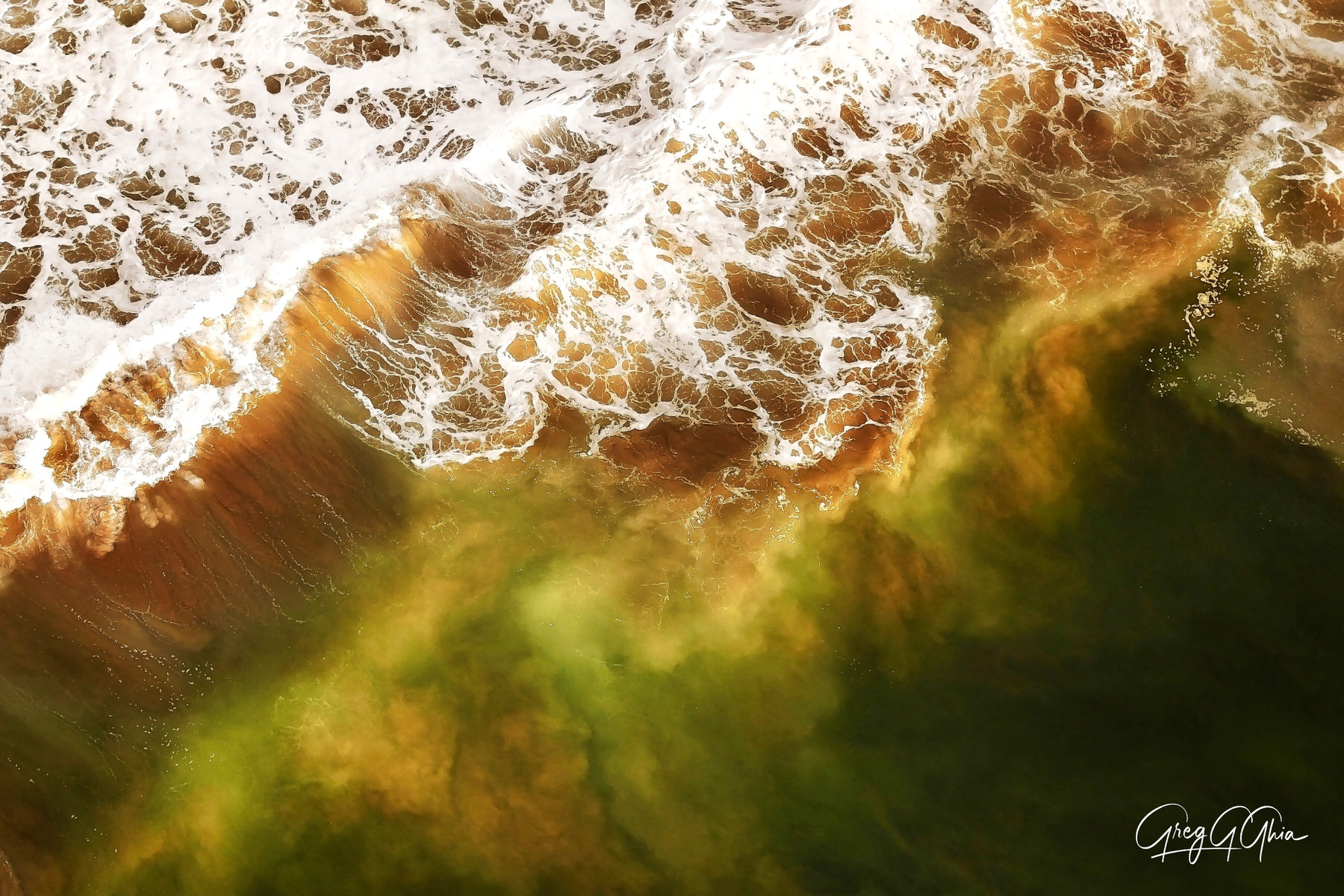 Aerial view with emerald green waves crashing into the coffee toned shore with white foam of the waves as they break onto the shore. 