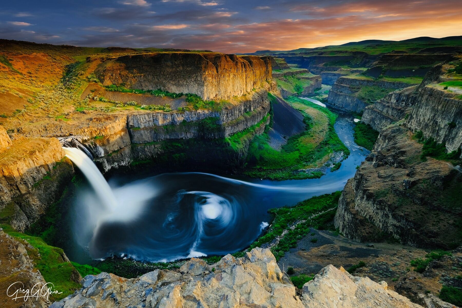 Waterfall tumbles into the swirling pool of water below the jagged cliffs while sunlight bathes the cliffs in light at sunset with pink clouds in the sky.